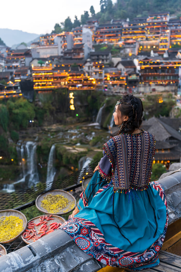 Mujer con vestido étnico tradicional observando desde un tejado el pueblo antiguo de Furong con sus cascadas e iluminación nocturna.