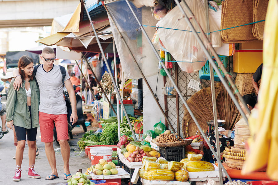 Una pareja de turistas paseando abrazados por un mercado local al aire libre en China, rodeados de puestos de frutas y verduras frescas.